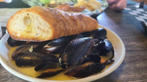 A bowl of mussels in broth, topped with a slice of toasted bread, served on a wooden table.