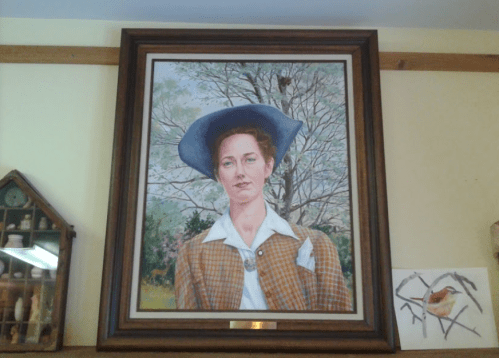 A framed portrait of a woman in a blue hat and brown checkered jacket, set against a nature background.