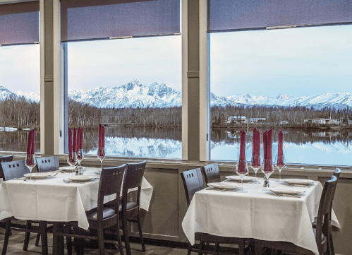 Elegant restaurant setting with white tablecloths, wine glasses, and a stunning mountain view through large windows.