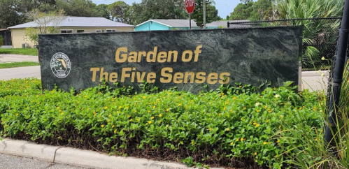 Sign for the "Garden of The Five Senses" surrounded by greenery and flowers, with a stop sign in the background.