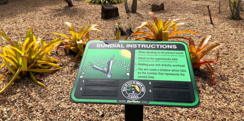 A sign with sundial instructions surrounded by gravel and plants, detailing how to read the time using the sundial.