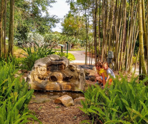 Two children play near a small stone waterfall surrounded by lush greenery and bamboo.