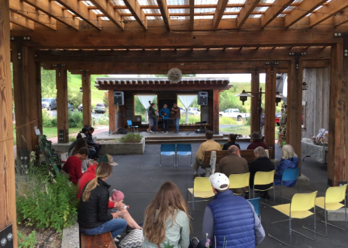 A small outdoor stage with musicians performing, surrounded by an audience seated on colorful chairs under a wooden structure.