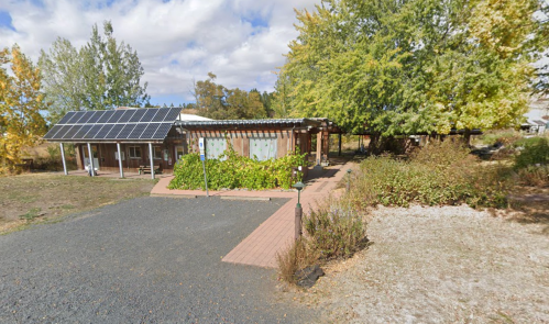 A small building with solar panels, surrounded by greenery and trees, under a partly cloudy sky.