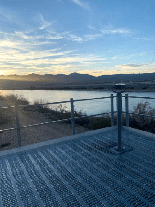 View from a lookout point with a metal railing, overlooking a river and mountains at sunset.