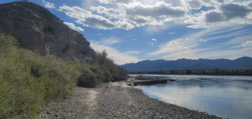 A serene riverbank scene with rocky shore, lush greenery, and mountains under a partly cloudy sky.