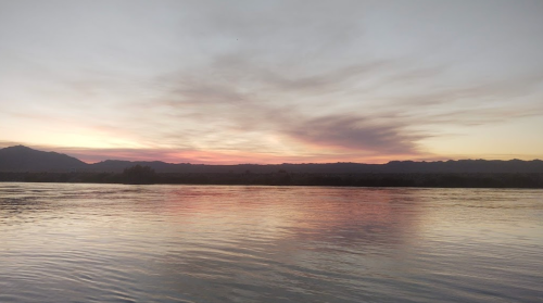 A serene river at sunset, reflecting soft pink and orange hues against a calm water surface and distant mountains.