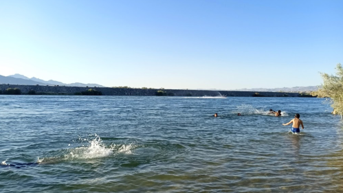 A person wades in shallow water near a river, with mountains in the background and others swimming in the distance.