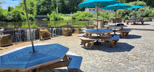 Outdoor seating area with wooden picnic tables and blue umbrellas by a river and waterfall. Lush greenery in the background.