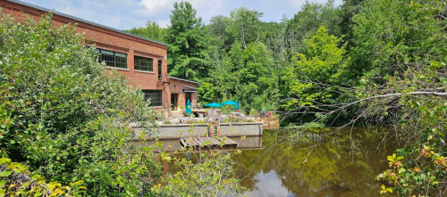 A serene view of a brick building by a calm pond, surrounded by lush greenery and outdoor seating with umbrellas.
