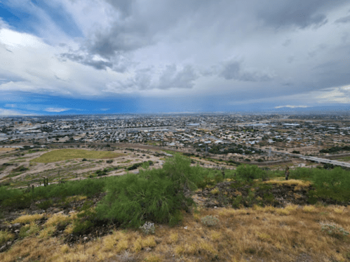 A panoramic view of a cityscape under a cloudy sky, with hills and greenery in the foreground.