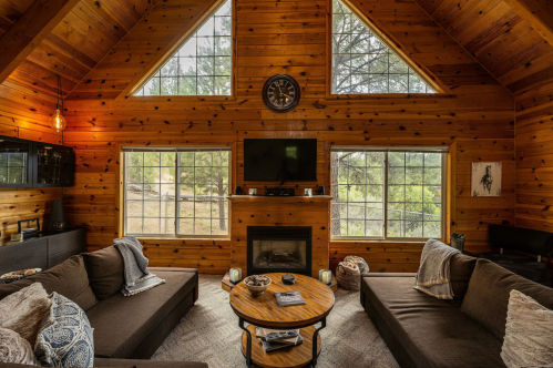Cozy wooden cabin interior with large windows, a fireplace, and comfortable seating arranged around a circular coffee table.