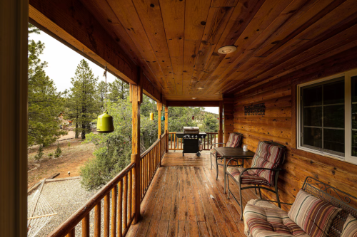 A wooden porch with a grill, chairs, and a table, surrounded by trees and a gravel path.