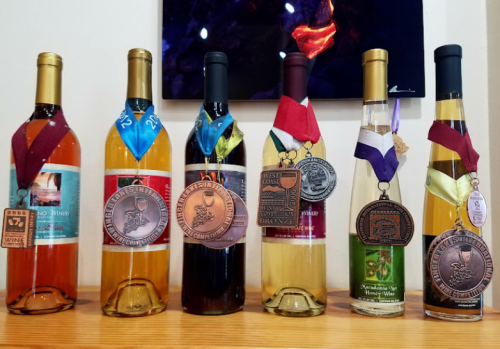 A display of six wine bottles, each adorned with medals and ribbons, showcasing various awards and achievements.