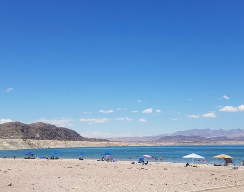 A sandy beach with umbrellas, people relaxing by a calm lake under a clear blue sky and distant mountains.
