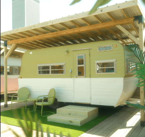 A vintage green and white trailer under a wooden canopy, with two chairs and a small wooden deck in front.