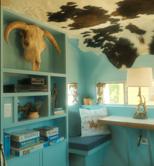 Cozy interior of a vintage trailer with turquoise walls, a cowhide ceiling, and rustic decor including a skull and plants.