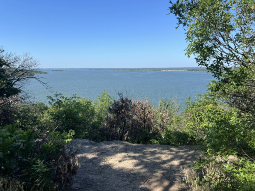 A serene view of a lake surrounded by greenery under a clear blue sky.