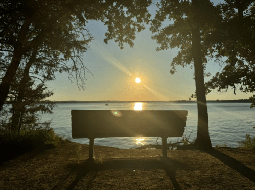 A wooden bench by a lake at sunset, with sunlight reflecting on the water and trees framing the scene.