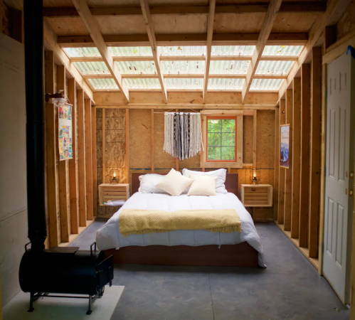 Cozy bedroom with a skylight, wooden walls, a bed with yellow accents, and a small wood stove.