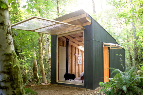 A modern cabin in a forest, featuring a large open entrance and a wood stove inside. Sunlight filters through the trees.