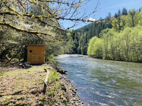 A serene river scene with a wooden cabin, surrounded by lush greenery and mountains under a clear blue sky.