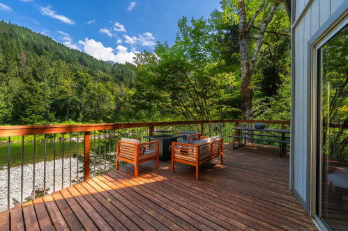 A wooden deck with two chairs overlooks a lush green landscape and a river, surrounded by trees and mountains.