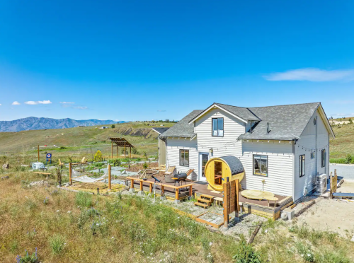 A modern house with a circular entrance, surrounded by a grassy landscape and mountains under a clear blue sky.