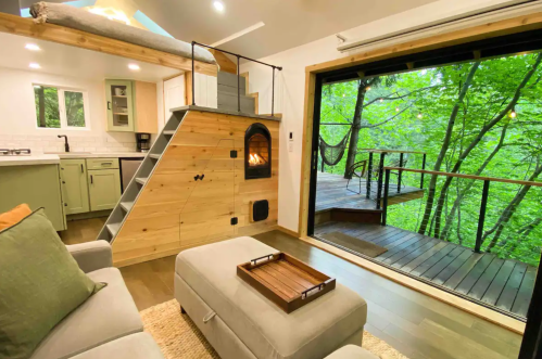 Cozy interior of a tiny home with a loft, fireplace, and large windows overlooking a lush green forest.