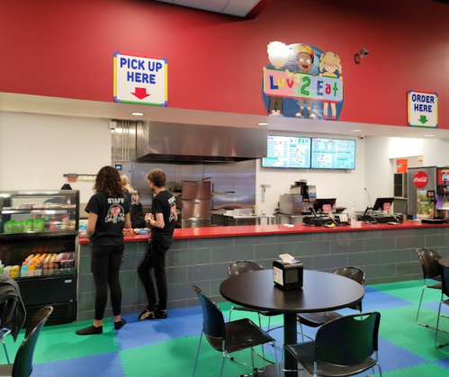 A colorful food counter with staff, menu boards, and seating area in a casual dining space. Signs indicate order and pickup.