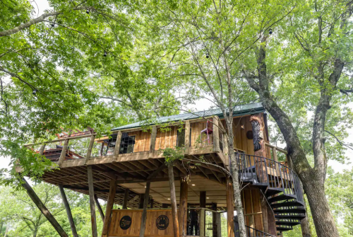 A wooden treehouse elevated among lush green trees, featuring a spiral staircase and a cozy balcony.