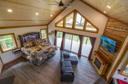 Cozy cabin interior with a large bed, seating area, and large windows overlooking nature. Warm wood accents throughout.