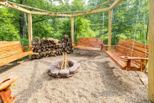 A cozy outdoor seating area with swinging benches around a fire pit, surrounded by greenery and stacked logs.