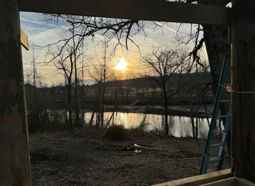 Sunset over a calm river, framed by trees and a wooden structure, with a ladder nearby.