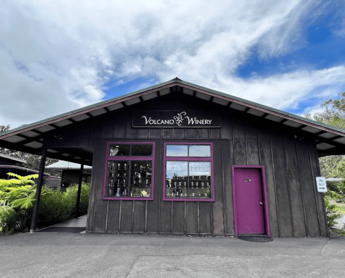 A wooden building with a sign reading "Volcano Winery," featuring large windows and a purple door, set against a cloudy sky.