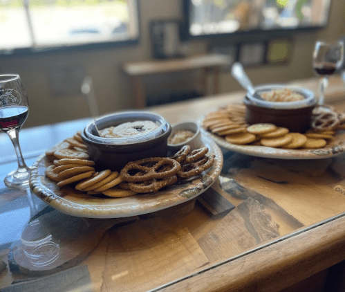 A wooden table with two platters of cookies, pretzels, and dips, alongside glasses of wine.