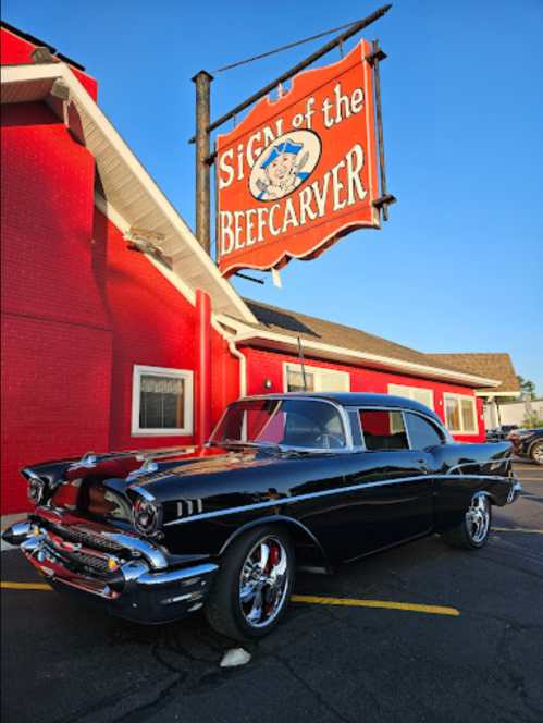 A classic black car parked in front of a red building with a sign that reads "Sign of the Beefcarver."