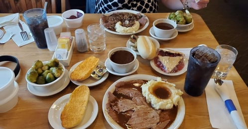 A table set with two plates of food, including meat, mashed potatoes, Brussels sprouts, bread rolls, and drinks.