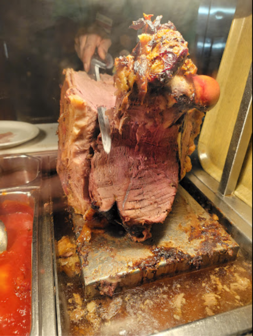 A large cut of meat on a carving station, with a hand holding a carving knife, surrounded by condiments.