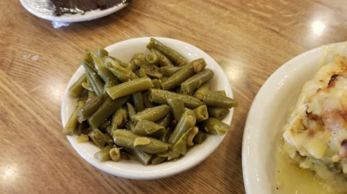 A small bowl of cooked green beans on a wooden table, next to a plate of food.