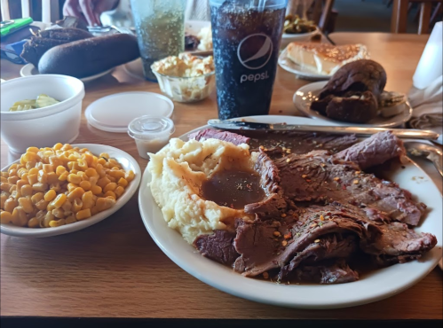A plate of sliced roast beef with gravy, mashed potatoes, corn, and drinks on a wooden table.