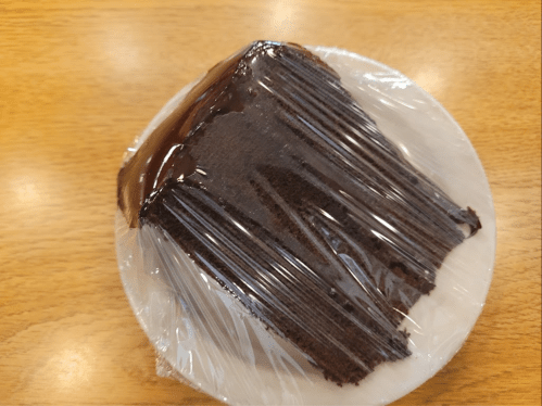 A slice of chocolate cake covered in plastic wrap on a white plate, resting on a wooden table.