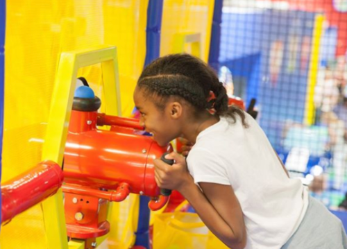 A girl with braided hair plays at a colorful indoor playground, interacting with a bright red toy.