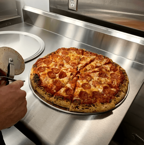 A freshly baked pepperoni pizza on a metal countertop, with a pizza cutter ready to slice it.