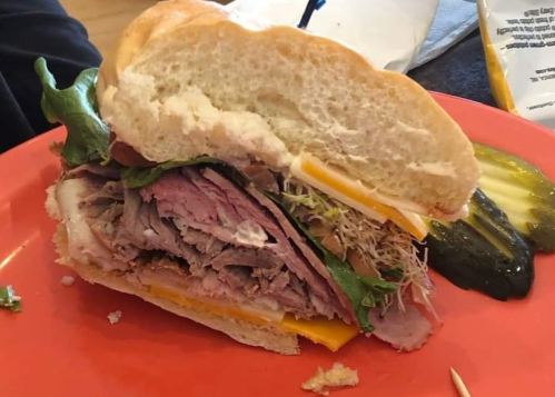 A close-up of a sandwich with layers of meat, cheese, lettuce, and sprouts on a plate, accompanied by a pickle.