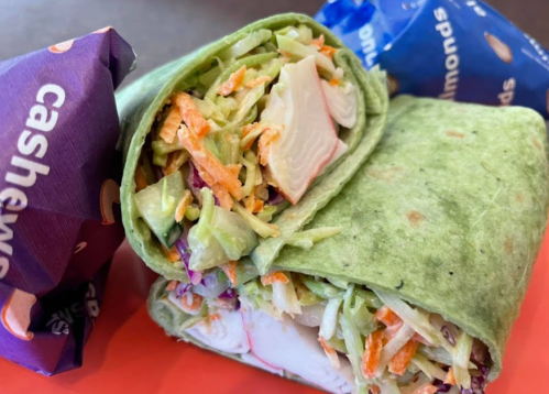 A green wrap filled with shredded vegetables, alongside two snack bags of cashews and pretzels on an orange plate.