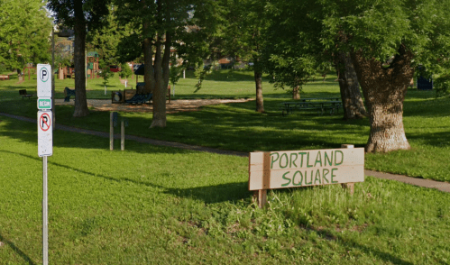 A park with green grass, trees, and a sign reading "Portland Square" near a parking sign.
