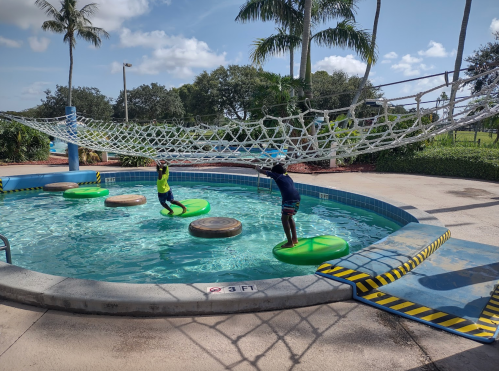 Two children play on floating platforms in a pool, with a net structure overhead and palm trees in the background.