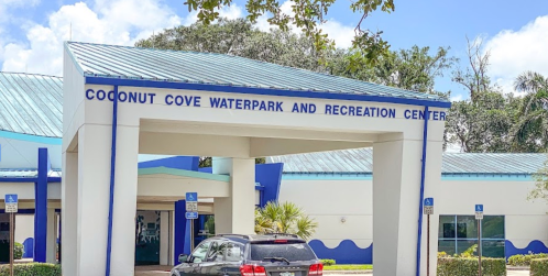 Entrance of Coconut Cove Waterpark and Recreation Center with a blue roof and palm trees in the background.