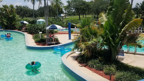 A lazy river pool with people floating on tubes, surrounded by palm trees and lush greenery on a sunny day.
